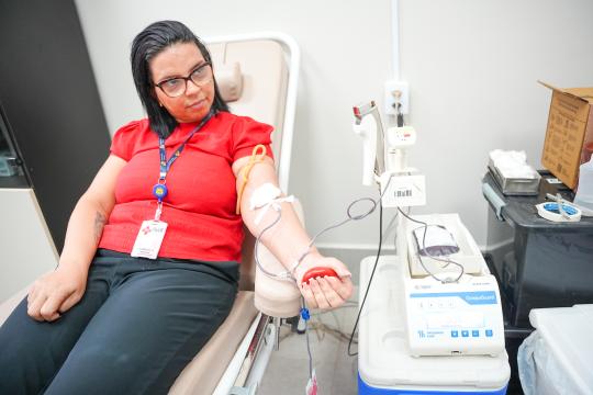 A imagem é uma fotografia de ambiente interno que retrata uma mulher sentada em uma poltrona reclinável branca durante um procedimento médico. Ela veste uma camiseta vermelha, calça jeans azul e óculos de grau, e possui um crachá pendurado no pescoço. Um acesso venoso está inserido em seu braço direito, conectado a uma máquina de hemodiálise branca com um visor digital que exibe informações numéricas. À direita da poltrona, há uma mesa com equipamentos médicos, incluindo um monitor, um frasco de soro e diversos dispositivos. Ao fundo, à esquerda, observa-se uma parede branca e, à direita, uma estante com caixas empilhadas. Uma tomada elétrica branca é visível na parede, acima da máquina.
