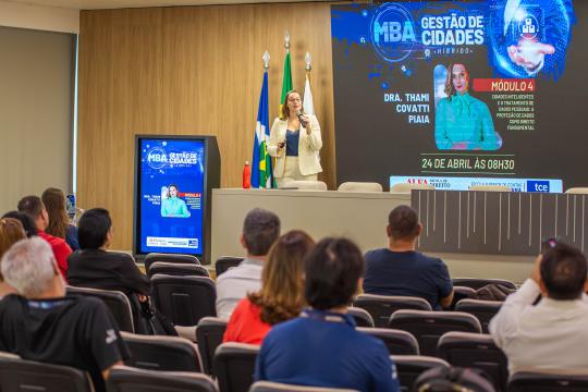 A imagem é uma fotografia tirada em um auditório durante um evento sobre MBA em Gestão de Cidades. No centro, uma mulher de cabelos castanhos e óculos, vestindo um blazer bege, está em um palco, com um microfone à sua frente e um banner atrás com os dizeres