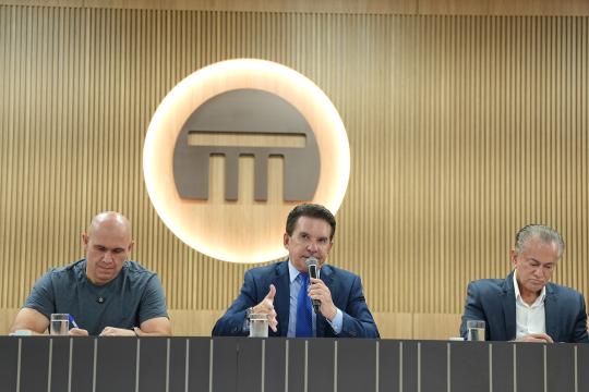 A imagem é uma fotografia que mostra três homens sentados em uma mesa durante uma reunião ou evento formal. À esquerda, Abilio Brunini prefeito de Cuiabá-MT, vestindo uma camiseta cinza, olha para a esquerda. No centro, Conselheiro Sérgio Ricardo presidente do Tribunal de contas de MT de terno azul e gravata, está falando em um microfone com a mão direita em gesto de fala. À direita, Desembargador Orlando Perry , com cabelos grisalhos, veste um blazer escuro e uma camisa clara, olhando para frente. Atrás deles, na parede, há um painel de madeira vertical e, ao centro, um círculo branco com um ícone dourado que representa uma construção com colunas. Uma mesa escura com garrafas de água e microfones está à frente dos homens, separando-os do observador.