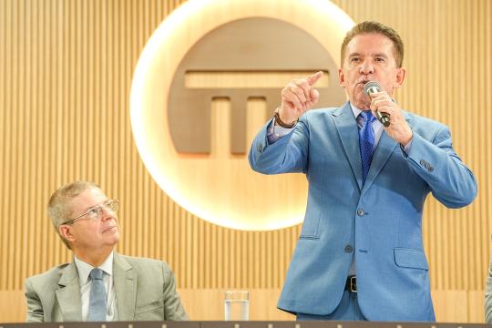 A imagem é um retrato em close-up de dois homens em um ambiente formal. À esquerda, um homem mais velho, de cabelos grisalhos e óculos, veste um terno cinza e uma gravata azul, observando atentamente. À direita, um homem de cabelos escuros e bem penteados, com um terno azul claro e gravata estampada, discursa em um microfone com a mão direita levantada em um gesto expressivo. Ao fundo, um grande círculo em tons de dourado e cinza exibe o símbolo de uma letra