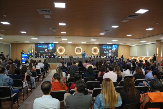 A imagem é uma fotografia de um evento formal realizado em um auditório. No centro, uma mesa comprida com uma toalha verde clara está disposta em frente a uma plateia composta por aproximadamente cinquenta pessoas sentadas em cadeiras pretas dispostas em fileiras. Atrás da mesa, há um painel com o logotipo