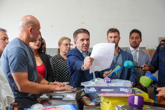 A imagem é uma fotografia jornalística que retrata um grupo de homens e mulheres em um ambiente interno. À esquerda, um homem careca com camisa azul e uma mulher com blusa estampada e óculos observam atentamente um homem de terno azul que está no centro da imagem, gesticulando com um documento na mão e falando. À direita desse homem, há mais dois homens, um com barba e outro sem, ambos com semblante sério e microfones próximos aos rostos. Em frente ao grupo, uma mesa de madeira exibe diversas caixas de presente coloridas, algumas abertas mostrando brinquedos. Ao fundo, parte de uma porta branca e um corredor são visíveis.