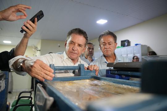 Fotografia em ambiente interno mostra dois homens em primeiro plano e outras pessoas ao fundo. À esquerda, um homem de camisa azul e branca segura um celular com a mão esquerda, enquanto aponta com a mão direita para algo fora do campo de visão. No centro, um homem de cabelos grisalhos e camisa branca observa atentamente um equipamento azul com duas alças, que está sobre uma superfície. À direita desse homem, outro indivíduo com camisa branca e gravata observa a mesma cena. Ao fundo, há mais pessoas, incluindo uma mulher sentada com um tablet, em um ambiente que parece ser um escritório ou local de trabalho, com teto branco e luzes fluorescentes.