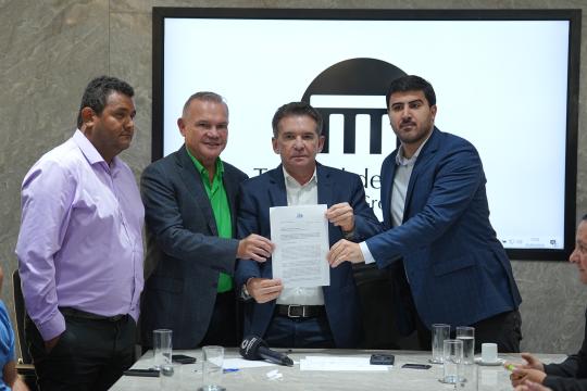 Fotografia em ambiente interno mostra quatro homens de terno em primeiro plano, em momento de assinatura de um documento. Da esquerda para a direita, o primeiro homem é de pele escura, camisa rosa e terno preto. O segundo homem é de pele clara, camisa verde e terno preto. O terceiro homem, ao centro, é de pele clara, camisa branca e terno azul marinho, e segura um documento em suas mãos, com outros dois homens apoiando as mãos sobre o papel. O quarto homem, à direita, é de pele clara, camisa branca e terno preto. Uma mesa de madeira clara está presente na frente dos homens, com dois microfones, copos de água e um tablet sobre ela. Ao fundo, há uma parede com textura de pedra e uma tela com um logotipo e informações sobre o evento com data '25/05/2023'.