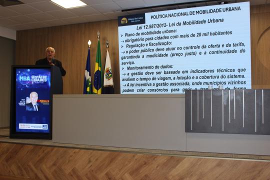 A imagem é uma fotografia de um evento ou palestra. No centro, um homem de terno e gravata gesticula enquanto fala, posicionado em frente a uma tribuna com um painel digital. O painel exibe o título 'POLÍTICA NACIONAL DE MOBILIDADE URBANA' e o texto 'Lei 12.587/2012– Lei da Mobilidade Urbana' seguido de tópicos sobre plano de mobilidade urbana, obrigatoriedade para cidades com mais de 20 mil habitantes, regulação e fiscalização, e monitoramento de dados. À direita da tribuna, há as bandeiras do Brasil e do estado de Goiás. Em frente à tribuna, um púlpito com o logotipo 'GESTÃO DE CIDADES' e uma imagem de um homem, possivelmente o palestrante, com a legenda 'Prof. Dr. Luiz Alberto Polonia'. O piso apresenta um padrão de mármore claro.