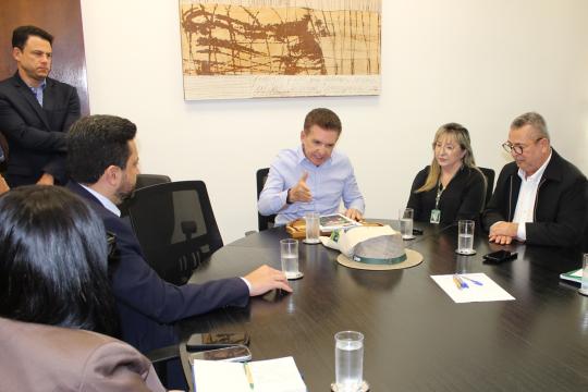Fotografia de um grupo de cinco pessoas em uma reunião formal. À esquerda, um homem de terno preto e camisa branca observa a cena. No centro, um homem de camisa azul e terno conversa gesticulando com as mãos, sentado à mesa. À direita do homem central, uma mulher com óculos e crachá observa atentamente, e ao lado dela, um homem de terno e cabelo grisalho também está presente. Em primeiro plano, um homem de costas, vestido com um terno preto, observa documentos sobre a mesa. Sobre a mesa, há papéis, canetas, um chapéu marrom, copos de água e telefones celulares. Ao fundo, na parede, há uma obra de arte abstrata em tons de marrom e bege.