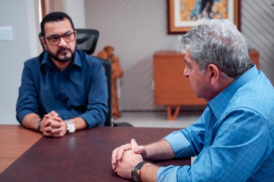 A imagem é um retrato de dois homens em um ambiente interno que aparenta ser um escritório. À esquerda, um homem de pele morena, cabelos pretos curtos e óculos de grau escuros veste uma camisa azul escura e está sentado à mesa com os braços cruzados, usando um relógio no pulso esquerdo. À direita, um homem de pele clara e cabelos grisalhos curtos usa uma camisa azul com padronagem quadriculada e está sentado de frente para o outro, com as mãos juntas sobre a mesa e um relógio no pulso esquerdo. Entre os homens, ao fundo, há uma cadeira de escritório preta e um aparador marrom com um quadro na parede acima dele, o qual exibe uma imagem com tons quentes. A iluminação do ambiente é suave e o foco está nos dois homens.