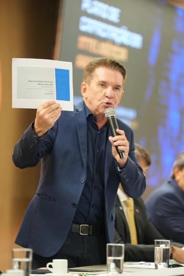 Fotografia de um homem, de pé, em um evento formal, segurando um documento e falando em um microfone. Ele veste um terno azul marinho com camisa clara, possui cabelos loiros penteados para trás e rosto com traços marcantes. O documento que ele segura exibe o título 'Plano Estratégico de Estado Mato Grosso 2050' e a data '13 de abril de 2023'. Ao fundo, uma parede com grafismos em tons de azul e dourado, e uma mesa com copos de água, xícaras e outras pessoas sentadas, em segundo plano e parcialmente visíveis.