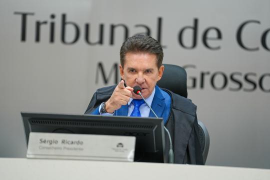 A imagem é um retrato de um homem de meia idade, com cabelos grisalhos e curtos, vestindo um terno azul marinho e gravata estampada, discursando em um microfone. Ele está sentado à frente de uma mesa com um computador portátil da marca Acer e uma identificação com o nome 'Sergio Ricardo' e o cargo 'Corregedor Presidente'. Ao fundo, uma parede com a inscrição 'Tribunal de Contas' e 'Mato Grosso' em letras maiúsculas. O foco da imagem está no homem, que aparece em primeiro plano com expressão séria enquanto fala.