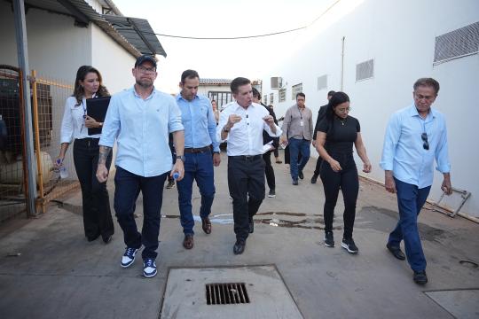 A imagem é uma fotografia colorida que retrata um grupo de pessoas caminhando em um espaço externo que parece ser um pátio ou área de serviço industrial. Do lado esquerdo, uma mulher de cabelos castanhos longos, vestindo camisa branca, calça preta e segurando uma prancheta, acompanha um homem barbudo com boné, camisa azul clara e jeans. Ao centro, seguem outros dois homens de camisa branca e calça social, um deles com uma pasta na mão. Mais adiante, uma mulher de cabelos escuros e camisa preta, com calça jeans, e um homem de camisa azul clara e calça jeans completam o grupo. Ao fundo, outros indivíduos estão visíveis, caminhando na mesma direção. O chão é de concreto, com uma grade de metal no primeiro plano. O cenário inclui paredes brancas e telhados de metal, sugerindo uma construção utilitária.