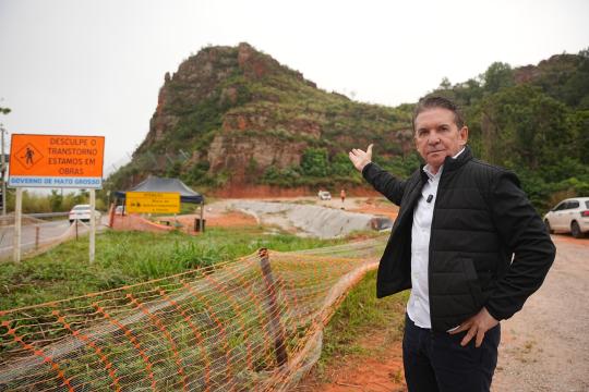 A imagem é uma fotografia de um homem de meia-idade, com cabelo castanho escuro e usando uma jaqueta azul escura sobre uma camisa clara, que está em pé e acena com a mão direita para a câmera. Ele se encontra em frente a uma área de construção com terra avermelhada, cercada por uma fita de segurança laranja. À esquerda, há uma placa de advertência amarela com a inscrição 'DESCULPE O TRANSTORNO ESTAMOS EM OBRAS, GOVERNO DE MATO GROSSO' e um aviso menor de 'ATENÇÃO'. Ao fundo, observa-se uma colina rochosa com vegetação esparsa e árvores, sob um céu claro. Um pequeno toldo azul está visível atrás da placa e ao lado da área de construção. Uma estrada de terra e um carro estão presentes na parte inferior direita da imagem.