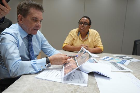 A imagem é um retrato de um homem de cabelos grisalhos e pele clara, vestindo uma camisa social azul clara com uma gravata azul escura, sentado à mesa e olhando para um documento aberto em suas mãos. Ele segura uma caneta azul e aponta para detalhes em uma fotografia colorida que faz parte do documento. À sua direita, em segundo plano, senta-se uma mulher de pele morena e cabelos presos, vestindo uma camisa amarela. Sobre a mesa, além do documento que o homem examina, há outros papéis com gráficos e anotações, e também outras fotografias de paisagens. Uma cadeira preta com encosto em tela está parcialmente visível à esquerda, e a mesa possui um tampo de madeira clara com veios aparentes. O fundo é uma parede lisa de cor clara.
