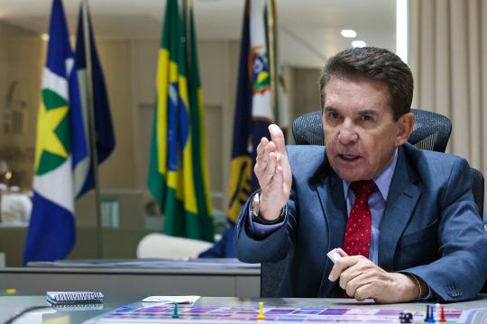 A imagem é um retrato de um homem, identificado como Arthur Lira, presidente da Câmara dos Deputados, sentado à mesa de reuniões. Ele veste um terno azul marinho, camisa branca e gravata vermelha com padrões florais, e gesticula com a mão direita aberta, enquanto aponta com o indicador da mão esquerda. Ao fundo, à esquerda, estão bandeiras do Brasil e de estados brasileiros, e à direita, uma luminária e parte de uma parede bege. A mesa em frente a ele é de madeira escura e contém papéis coloridos, canetas, um porta-canetas e um pequeno objeto vermelho retangular. O fundo é composto por um painel de madeira claro e uma janela que ilumina o ambiente.