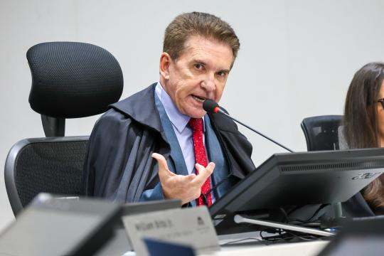 Fotografia de um homem de meia-idade, com cabelos castanhos e curtos, trajando um terno azul marinho, camisa branca e gravata vermelha estampada, sentado em uma cadeira de escritório preta com encosto alto. Ele está em primeiro plano, olhando para a câmera com uma expressão séria, gesticulando com a mão direita aberta e segurando um microfone com a mão esquerda. Ao fundo, à direita, parcialmente visível, encontra-se uma pessoa com cabelos escuros e um monitor de computador preto. Em frente ao homem há uma mesa com um pequeno suporte para placa de identificação e um laptop. A parede atrás dele é branca.