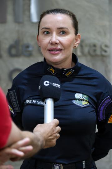 Fotografia de uma policial militar em primeiro plano, vestindo uniforme azul escuro com detalhes dourados, incluindo o brasão da Polícia Militar e uma identificação com o nome 'Ten Cel PM Ludmila'. Ela olha diretamente para a câmera com um leve sorriso, usando um microfone de lapela com o logotipo da 'TV Contas Canal 302'. Ao lado esquerdo dela, parcialmente visível, está a sigla 'PMMT' em letras vermelhas sobre fundo branco, e à direita, um emblema da 'Guarda Municipal de Mato Grosso do Sul'. No fundo, desfocado, aparece uma parede com inscrições em letras brancas e algumas pessoas. A policial usa brincos de argola dourados e uma pequena corrente com um pingente.