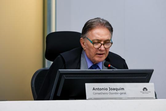 A imagem é um retrato de Antônio Joaquim, um homem idoso de cabelos grisalhos e curtos, usando óculos de armação fina e dourada. Ele está sentado em uma cadeira preta com encosto alto e usa uma camisa social branca com uma gravata azul. Um microfone está posicionado em frente a ele, e em sua frente há um monitor de computador preto ligado. Sobre uma mesa marrom clara, à frente do monitor, há uma identificação com o nome 'Antônio Joaquim' e abaixo, 'Conselheiro Ouvidor-geral', acompanhado do logo do Tribunal de Contas do Estado de Mato Grosso. O fundo é uma parede lisa de cor clara.