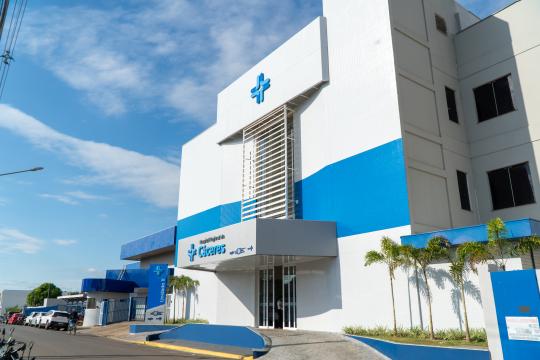 A imagem é uma fotografia de um edifício hospitalar moderno sob um céu azul claro com algumas nuvens brancas. O hospital possui uma fachada predominantemente branca com detalhes em azul, apresentando uma cruz azul no topo. Há uma sequência de janelas retangulares verticais com venezianas brancas no centro do edifício. Na parte inferior, a entrada do hospital é marcada por uma área coberta e portas de vidro, com o nome 'Hospital Regional de Cícero Dias' em letras azuis. À esquerda, há uma estrutura menor também em azul e branco, com a inscrição 'Unidade II'. Em frente ao hospital, há um estacionamento com alguns carros e motos, além de algumas palmeiras e árvores. Uma cerca azul com informações escritas e um portão de acesso estão à direita, completando a composição da imagem.