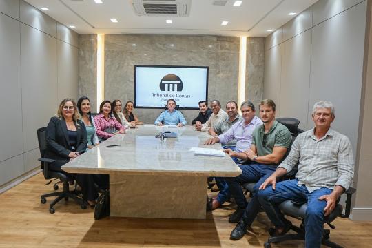 A imagem é uma fotografia de um grupo de pessoas reunidas em uma sala de conferências. No fundo, há uma parede com painéis texturizados em tons de marrom e um monitor exibindo o logo do 'Tribunal de Contas do Mato Grosso'. Uma longa mesa de madeira clara com superfície refletora ocupa o primeiro plano, ao redor da qual estão sentados dez homens e quatro mulheres. As pessoas estão vestidas de forma formal e casual, com ternos, blazers e camisas polo. Na extremidade central da mesa, um homem sentado em uma cadeira preta chama a atenção, e os demais membros do grupo estão distribuídos ao longo da mesa, alguns sentados e outros levemente inclinados para frente, olhando para a frente. No teto, há luminárias embutidas e um aparelho de ar condicionado. A sala possui paredes em tons neutros e o ambiente parece ser bem iluminado.